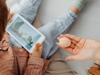 A child is absorbed in phone entertainment, with their parent seated beside them, holding a brain-shaped toy