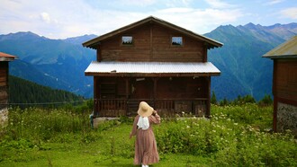 A woman wearing a hat walks toward a rustic wooden cottage surrounded by breathtaking mountain scenery and peaceful nature