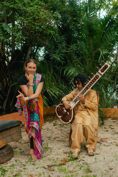 Amid a festive celebration at home, an Indian musician in traditional clothing plays the sitar outdoors, as a woman in a saree gracefully lights a diya lamp for Diwali