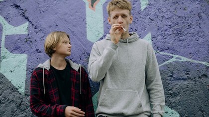 Two teenagers, one puffing on a cigarette while the other patiently waits for their turn