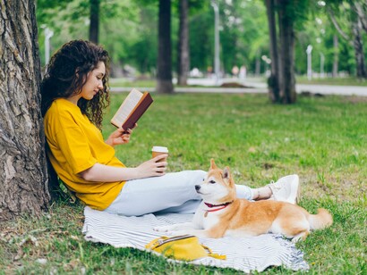 A young woman sitting beneath a tree and reading a book beside her dog, representing the restorative power of quiet leisure and time away from technology