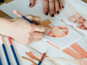 Uma mulher desenhando uma peça de roupa em uma mesa de trabalho. Desenhar moda é construir conceitos que irão dialogar com o mundo contemporâneo