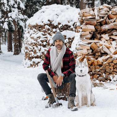 A man and his dog sitting beside a pile of firewood in the snow
