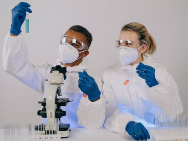 Pesquisadores equipados para o trabalho laboratorial realizam experimentos com tubos de ensaio e um microscópio. A pesquisa sobre o RNA mensageiro procura ampliar as possibilidades de tratamento do câncer, incluindo vacinas desenvolvidas para ajudar o organismo a reconhecer e combater células tumorais