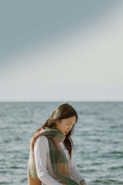 A peaceful scene of an Asian woman with long hair gazing at the ocean from the shoreline, capturing solitude and gentle loneliness