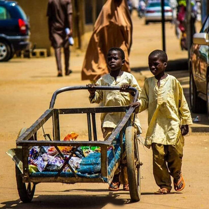 The picture shows two Almajiri children pushing a locally called "Push-push" cart, carrying a mix of goods for sale, including chocolate sweets, biscuits, chewing gum, and detergent