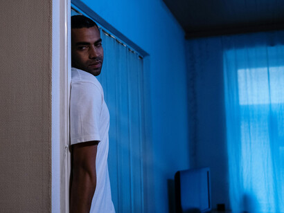 Man wearing a white T-shirt standing alone by a sliding door in a modern apartment with soft blue lighting