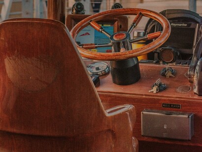 Cadeira diante da roda do leme. Estava sentada na ponta inicial do barco, se eu esticasse o braço um pouco, conseguiria tocar o volante, “volante”? 