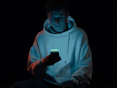 Sentado em um ambiente de pouca luz, um homem de moletom branco utiliza o celular, cuja luminosidade se destaca contra o fundo escuro. A composição concentra a atenção no usuário e no aparelho, deixando o restante do ambiente em segundo plano. Estamos usando o celular por interesse genuíno ou apenas para preencher o tempo e evitar o desconforto do tédio? O artigo convida a questionar esse comportamento