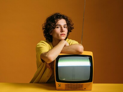 O jovem, vestido de amarelo, apoia os braços sobre um televisor antigo cuja tela apresenta uma faixa luminosa de interferência. A imagem remete ao lugar que a televisão ocupou na formação cultural de diferentes gerações, tema recuperado pelo autor ao recordar o período das aulas televisionadas e da difusão de conhecimentos pelos meios de comunicação