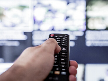 El mando apunta hacia una pantalla cargada de contenidos, símbolo de una audiencia que selecciona, cambia y consume información a un ritmo cada vez más inmediato