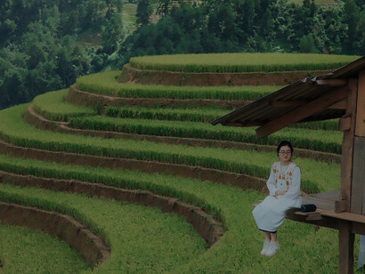 Amidst vast layers of agricultural land, a small hut stands as a symbol of sustainable farming practices. On its veranda, a woman sits, embodying the ideals of clean, wholesome food production