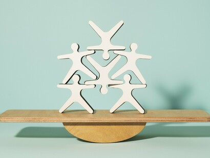Six abstract figures form a collaborative pyramid on a balancing board, representing many organizations' challenges when implementing Agile, like the need for a balance between structure and flexibility and planning and adaptation