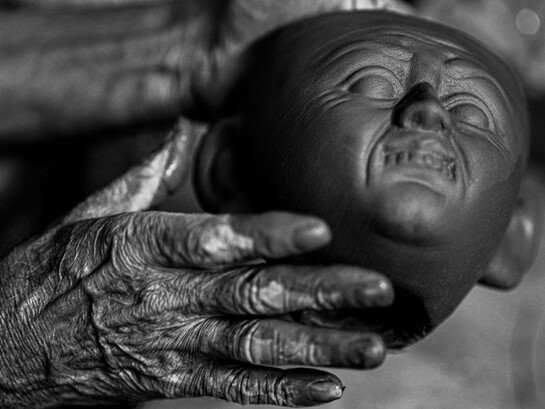 Um artesã idosa modela uma cabeça em argila, trabalhando cuidadosamente os volumes e os detalhes da escultura. Em suas mãos, a matéria ganha forma por meio de gestos que exigem prática, sensibilidade e paciência. Assim como no acordelado, a cerâmica artesanal preserva conhecimentos transmitidos pela experiência e transforma o trabalho manual em expressão artística