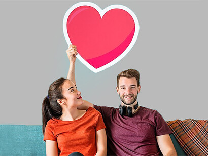 A cheerful couple sits on a couch, holding heart icons to represent red and green flags, showcasing romantic attraction and relationship dynamics