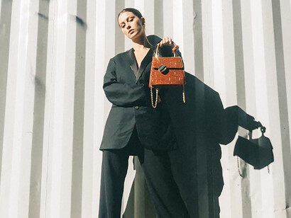 A woman wearing a black blazer and holding a brown handbag, leaning against a white wall, embodying old money fashion, generational wealth style, and classic preppy fashion