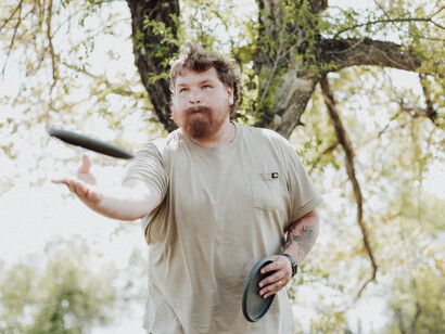 A man in mid-throw, sending a disc flying toward the disc golf basket
