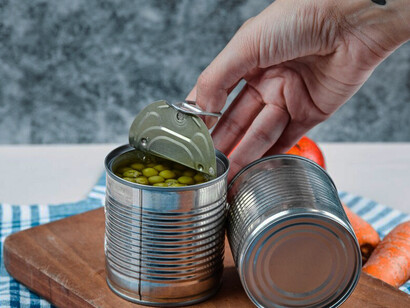 A hand holding a spoon reaches for two cans of boiled green peas placed on a vibrant tablecloth