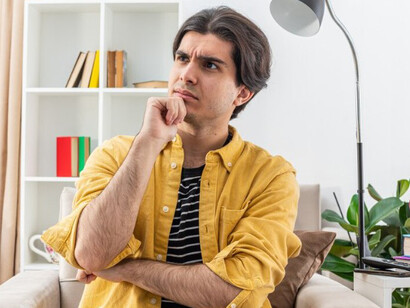 A casually dressed young man sits on a chair, looking aside thoughtfully with his hand on his chin