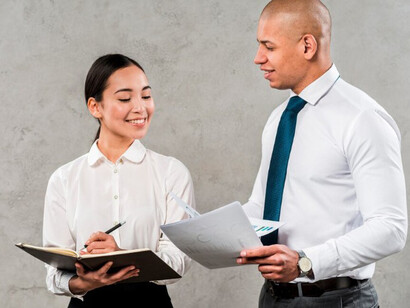A young businessman and businesswoman stand against a grey wall, deep in discussion over a project — a quiet snapshot of early-career ambition