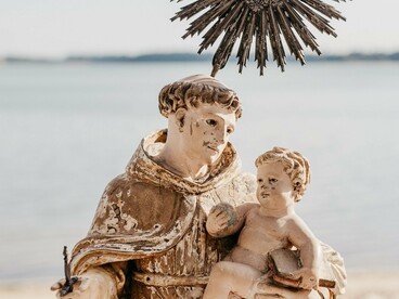 Em um enquadramento mais próximo, Santo Antônio aparece segurando o Menino Jesus, enquanto o mar compõe o cenário ao fundo. No romance A Cabeça do Santo, a devoção ao santo casamenteiro é transformada em elemento de uma história marcada por fé, desejos e encontros