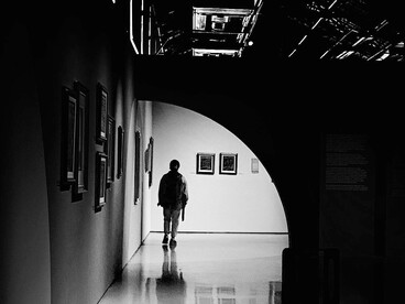 A dramatic black-and-white portrait of a lone figure in a vast empty hallway, where minimalist space and shadow evoke silence, isolation, and deep contemplation