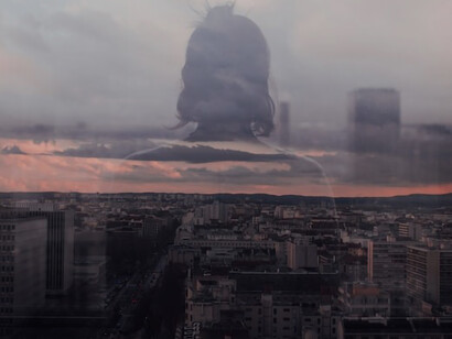 A woman's reflection, poised before a glass panel, contemplating the high-rise and mid-rise buildings