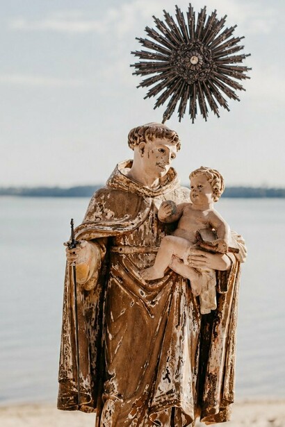 A representação de Santo Antônio ganha destaque diante da paisagem marítima, unindo a imagem religiosa ao ambiente natural. Na obra de Socorro Acioli, a figura do santo também ultrapassa o espaço da devoção e se torna parte do imaginário que dá vida à fictícia Candeia