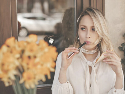 A stylish blonde woman in a white blouse, accessorized with pearl jewelry, smiles brightly while seated at a charming street café, gazing happily into the distance