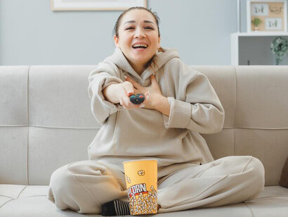 Relaxing on the couch in comfy clothes, a woman enjoys a cookie, remote in hand, while watching her favorite K-drama