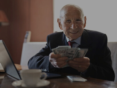 A contented senior businessman, seated at his desk, multitasking with a laptop and counting money