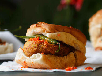 Restaurant scene showcasing a plate of Mumbai Vada Pav—deep-fried potato dumpling in a bread bun, garnished with green chili and tomato. In the foreground, an unrecognizable person reaches for a slice of tomato, with the plate in sharp focus