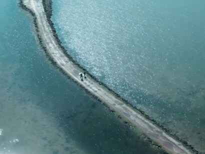 Two figures crossing a narrow causeway surrounded by open water, representing the precarious divide between safety and annihilation