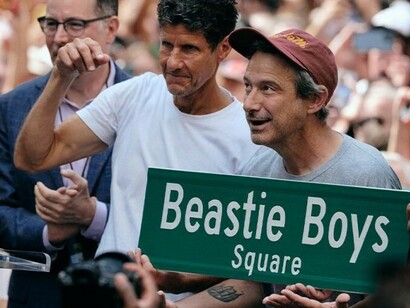 Um close-up do momento icônico: Ad-Rock (Adam Horovitz) e o vereador Lincoln Restler apresentam orgulhosamente a nova placa da 'Beastie Boys Square' para a multidão