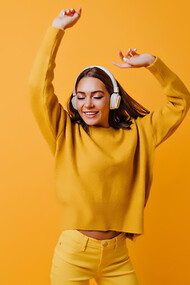 A carefree brunette woman in stylish yellow pants joyfully dancing to a good song, against a backdrop of an inviting orange wall, exuding warmth and happiness