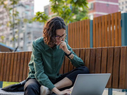 A young woman sits on a park bench, smiling as she joins a remote work video call on her laptop