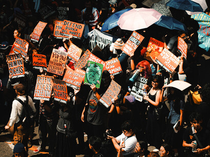 Vista cenital de una concurrida marcha de protesta con sombrillas y multitud de carteles reivindicativos como "Defend Press Freedom" / "Defender la libertad de prensa" y "Defend Human Rights" / "Defender los derechos humanos"