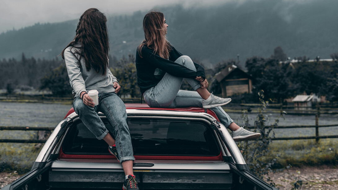 Amigas estacionam carro na estrada e sobem em cima do capô para adimirar a vista para as montanhas enquanto sonham com seus projetos pessoais