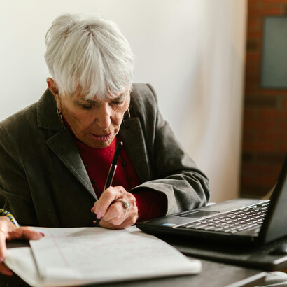 An elderly woman in a gray blazer working on her laptop at a café, representing the modern reality of global aging and lifelong productivity