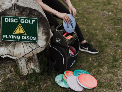 A person rests on a tree stump, surrounded by a backpack and a collection of disc golf discs