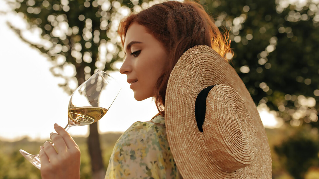 Stylish young girl with a foxy hairstyle donning a modern straw hat and trendy summer outfit poses elegantly with a glass of white wine amidst the picturesque vineyards of Hungary