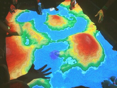 Desenvolvedores da Wikimedia no Ars Electronica Center em Linz, Áustria, brincando com um mapa topográfico em caixa de areia de realidade aumentada. A RA é uma ferramenta única para contextualizar conceitos abstratos e aproximá-los da realidade das crianças. Por exemplo, ao estudar o sistema solar, as crianças podem visualizar planetas em 3D, explorando suas características de maneira dinâmica e divertida