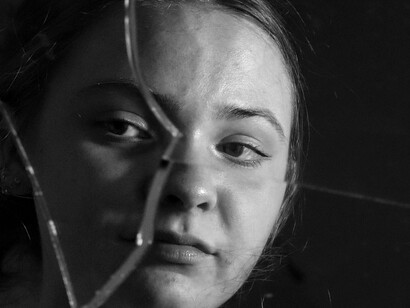 Mulher observa seu reflexo em espelho quebrado. Corremos, exibimos, acumulamos… mas raramente habitamos. Faltam verbos de ligação: ser, permanecer, tornar-se, ficar. Vivemos como quem está sempre de passagem — e o Naikan é uma forma de voltar. De pousar dentro da própria existência como quem retorna de uma longa viagem e precisa desfazer as malas com cuidado