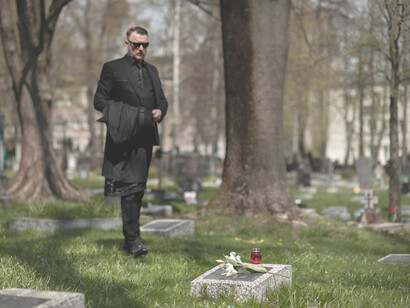 Mourning in a graveyard, a man reflects by a gravesite while a solemn funeral procession unfolds nearby