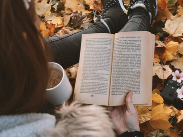 A person reading a book and holding a cup of coffee, surrounded by autumn leaves