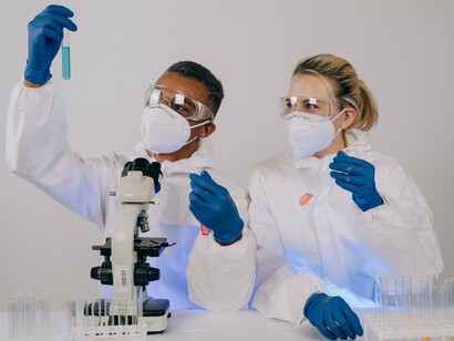 Pesquisadores equipados para o trabalho laboratorial realizam experimentos com tubos de ensaio e um microscópio. A pesquisa sobre o RNA mensageiro procura ampliar as possibilidades de tratamento do câncer, incluindo vacinas desenvolvidas para ajudar o organismo a reconhecer e combater células tumorais