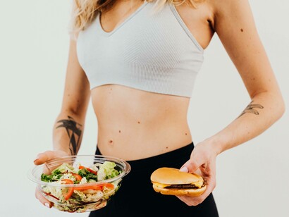 A woman in her training outfit holding a salad in one hand and a cheeseburger in the other