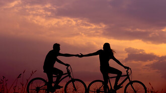 A woman on a bike reaching out to take the hand of a man riding behind her, both enjoying a moment of connection
