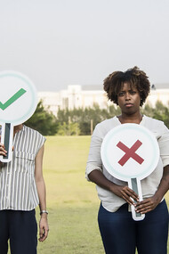 A dissatisfied Black woman gazes directly at the camera, holding a sign shaped like a cross