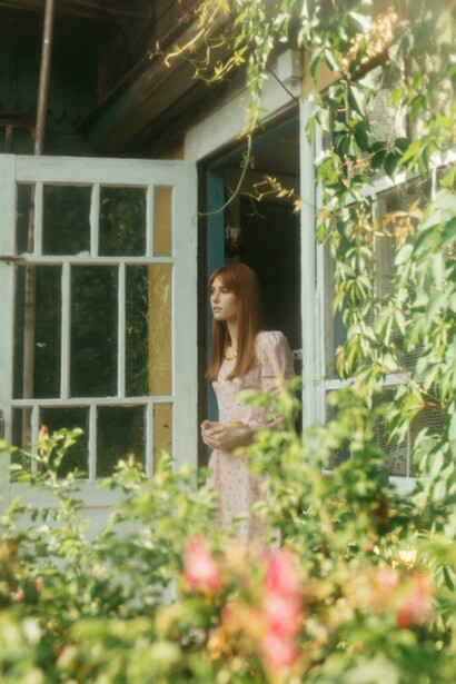 A young woman in a vintage dress stands at the entrance of a rustic country house, surrounded by lush greenery, blooming flowers, and soft summer light